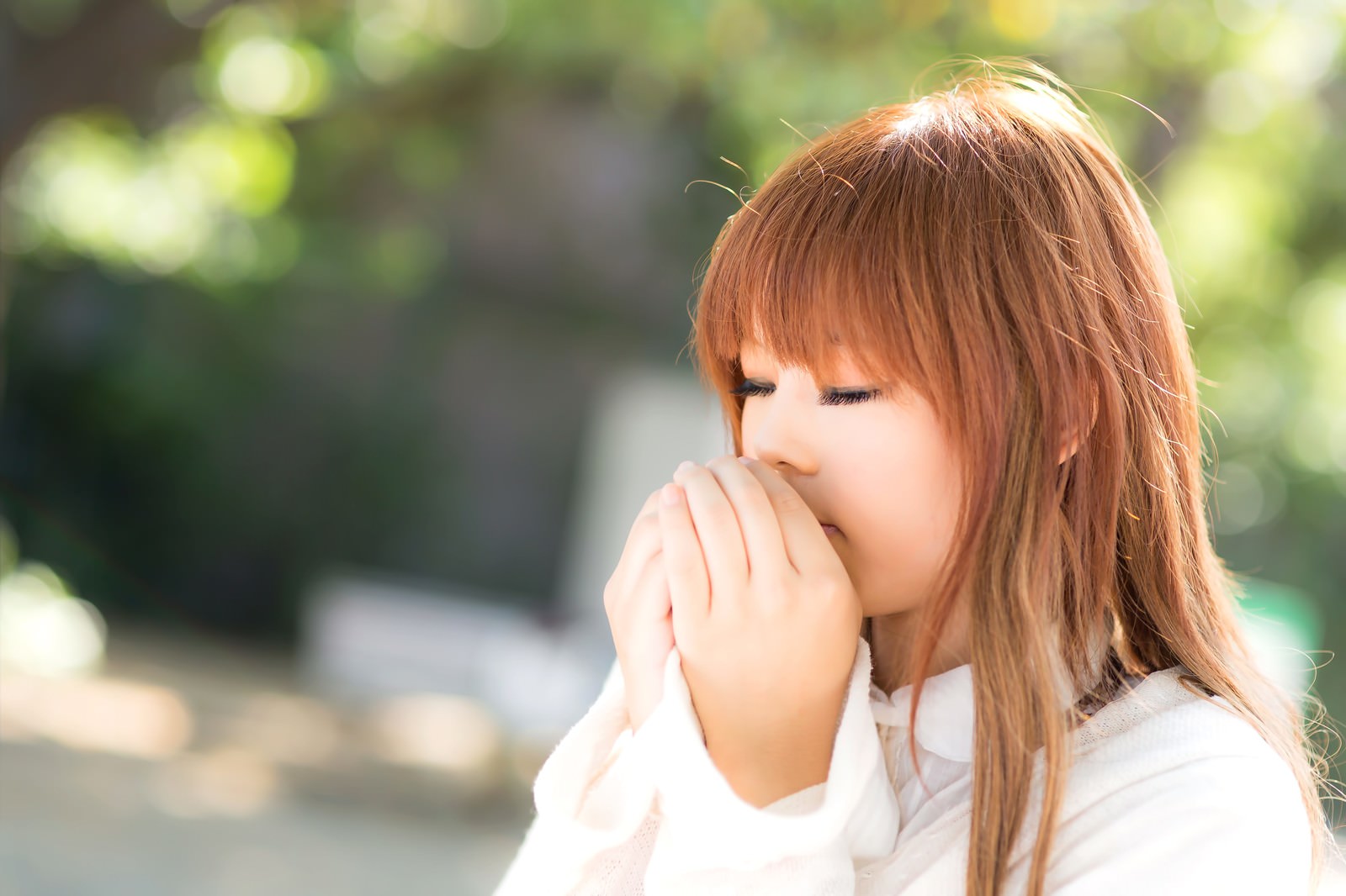 両手を口に寄せて吐息で温める、茶色い髪の女性が冬の屋外で口元を温めている［モデル：あみ］