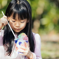虹色のかき氷をストローで飲む女の子の夏の風景の写真