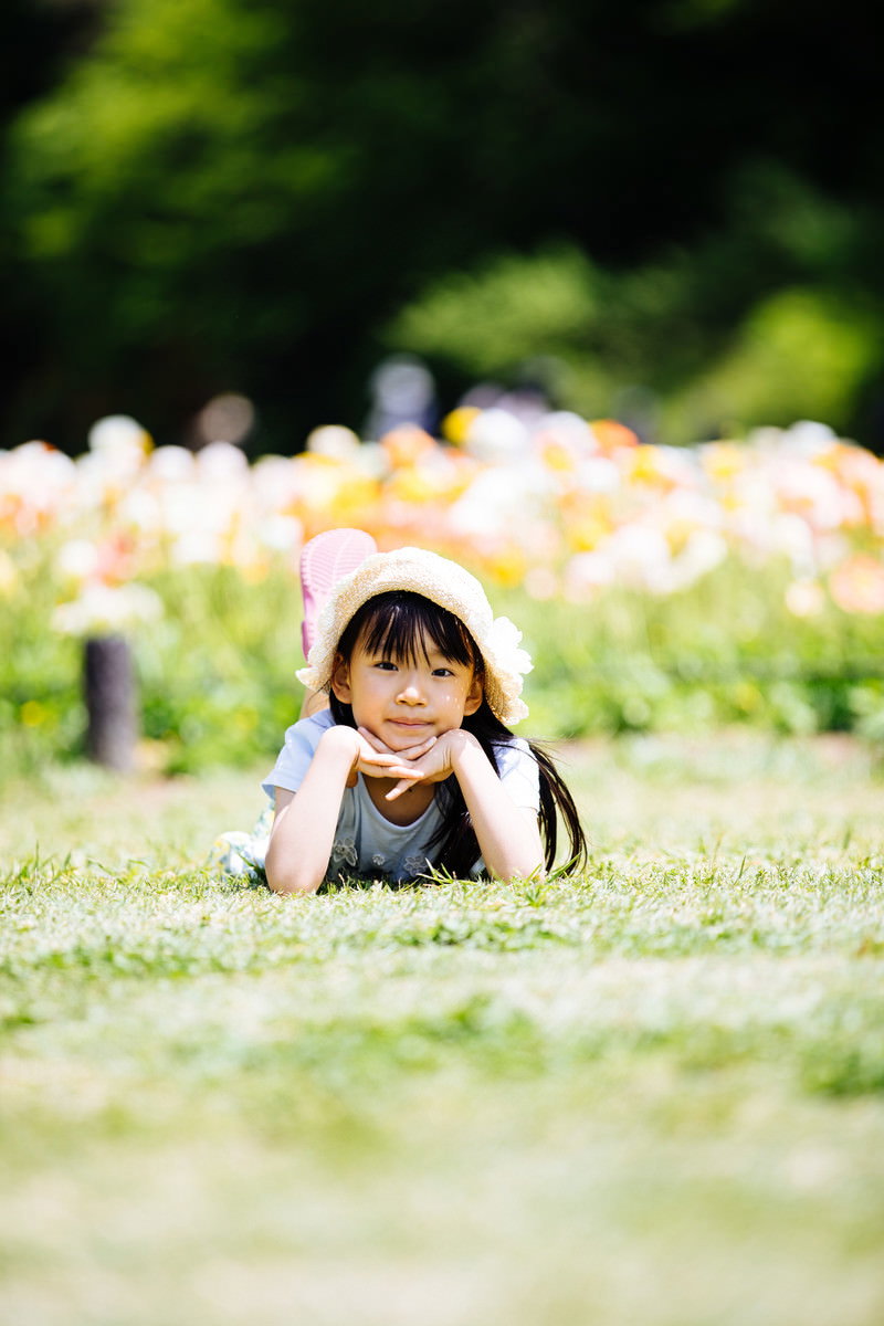 花畑を背景に芝生の上で顎をついてくつろぐ麦わら帽子の女の子［モデル：あんじゅ］