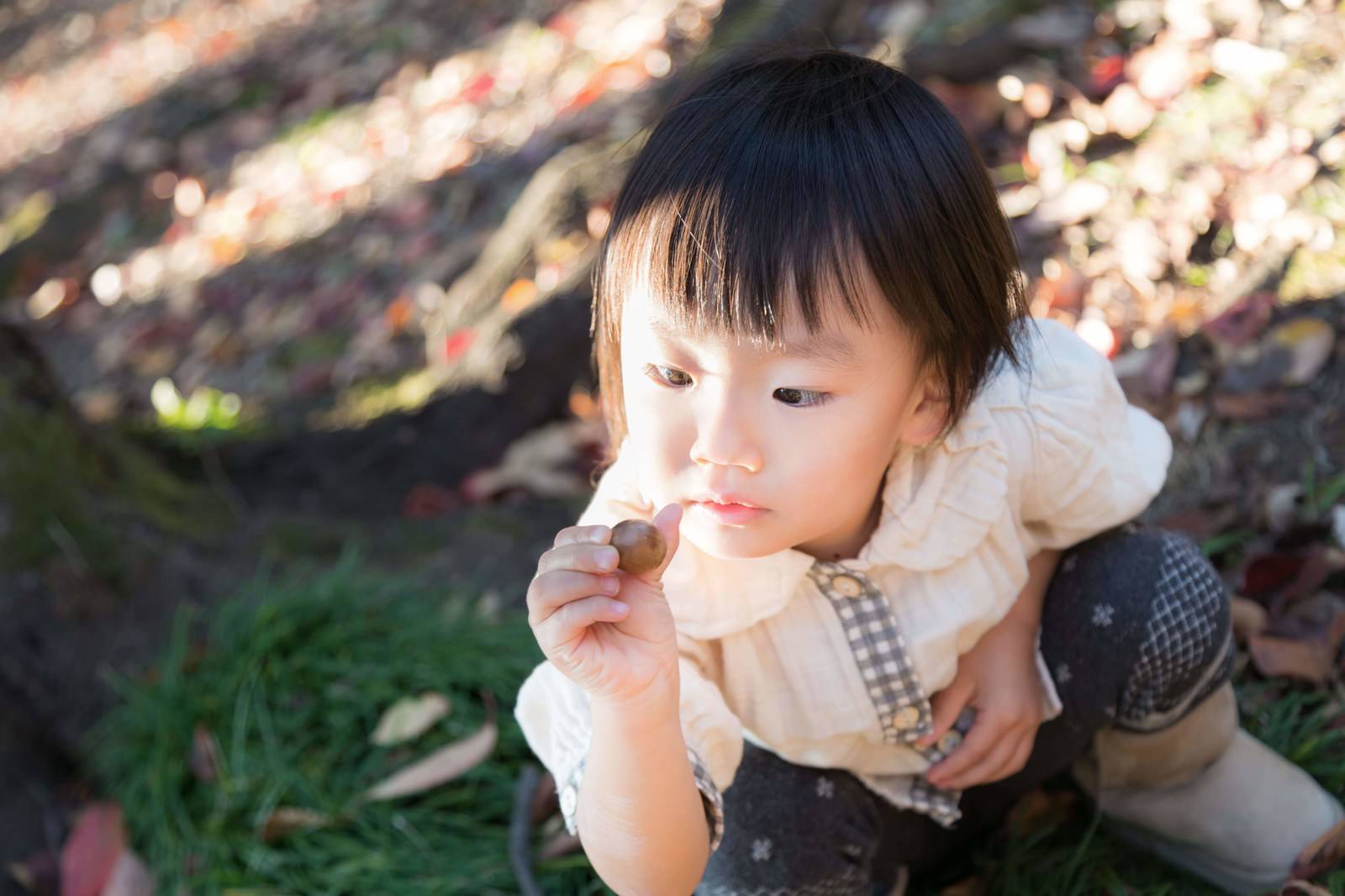 屋外の落ち葉の上で口元に手を当てて座る格子柄の幼児［モデル：あんじゅ］