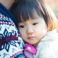 大人の肩に抱かれてピンクの物を持つ白い服の黒髪の幼児の写真