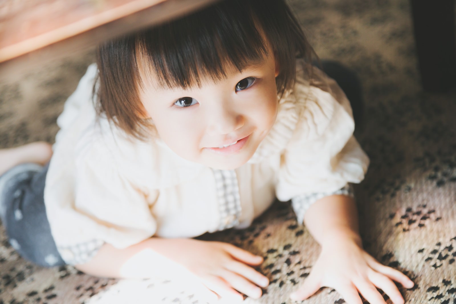 模様のある敷物の上で床に座る白い服の黒髪の子供［モデル：あんじゅ］