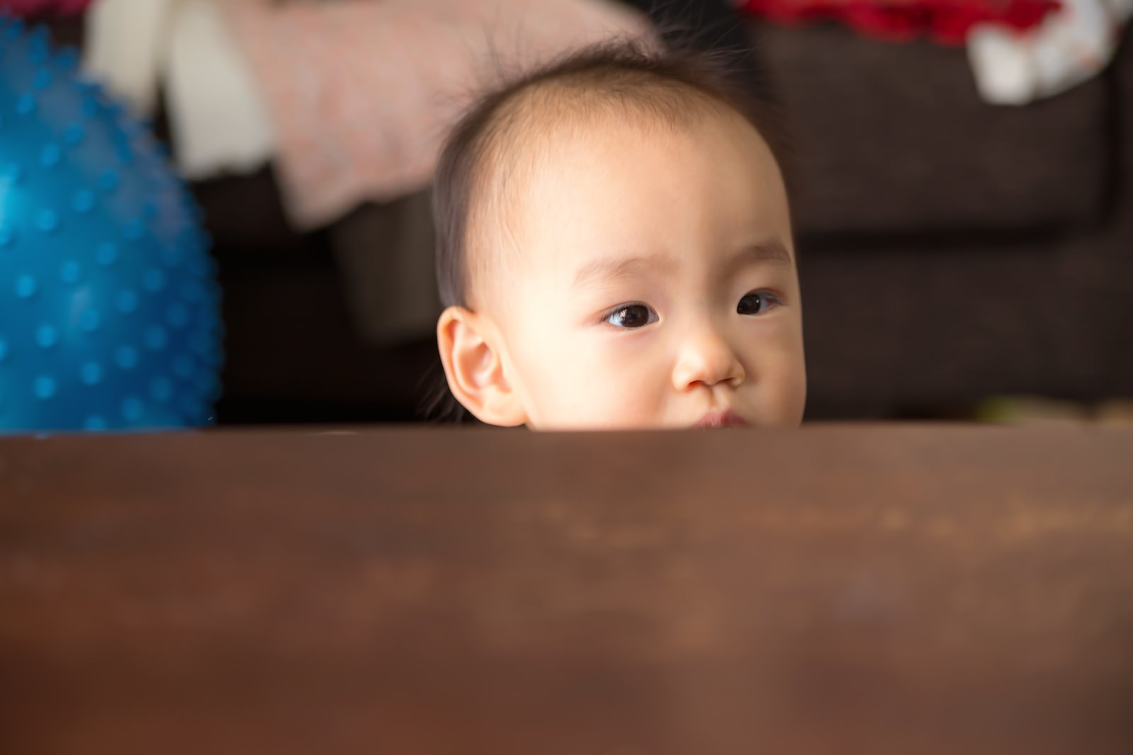 テーブルの端から顔を出し、瞳を開いて見つめる赤ちゃん［モデル：あんじゅ］