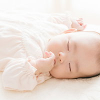 指を咥えて眠る新生児、白いベビー服姿の寝顔の写真