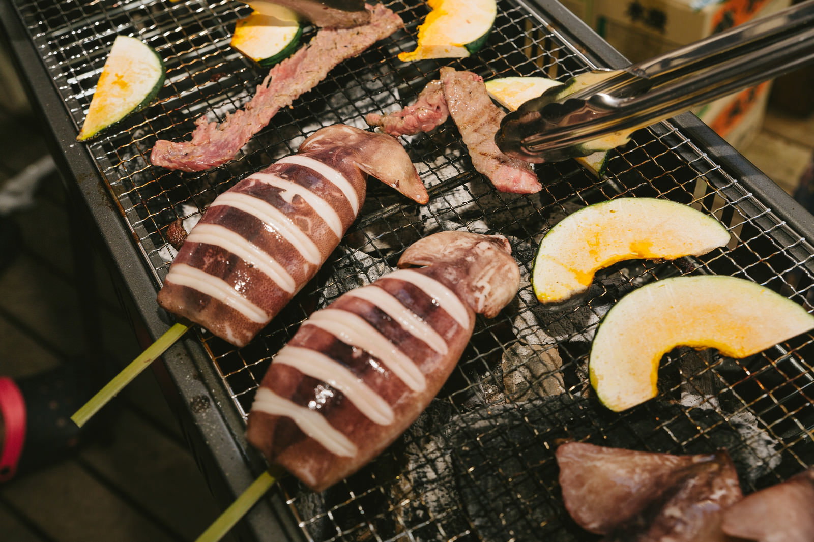 バーベキューで牛肉とイカをトングで焼く場面