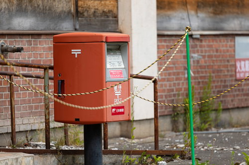 朽ちた廃墟の前に立つ赤い郵便ポスト、チェーンで囲まれた田舎の風景