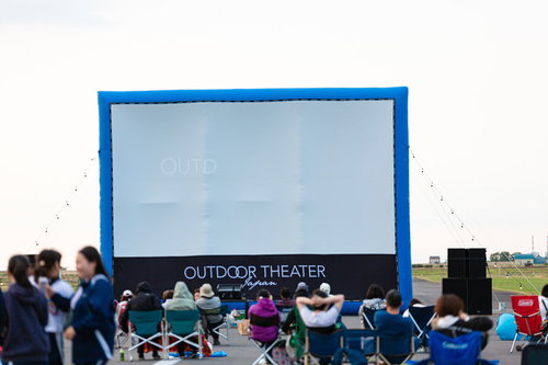 青いフレームの巨大スクリーンを前にした野外映画の上映前風景