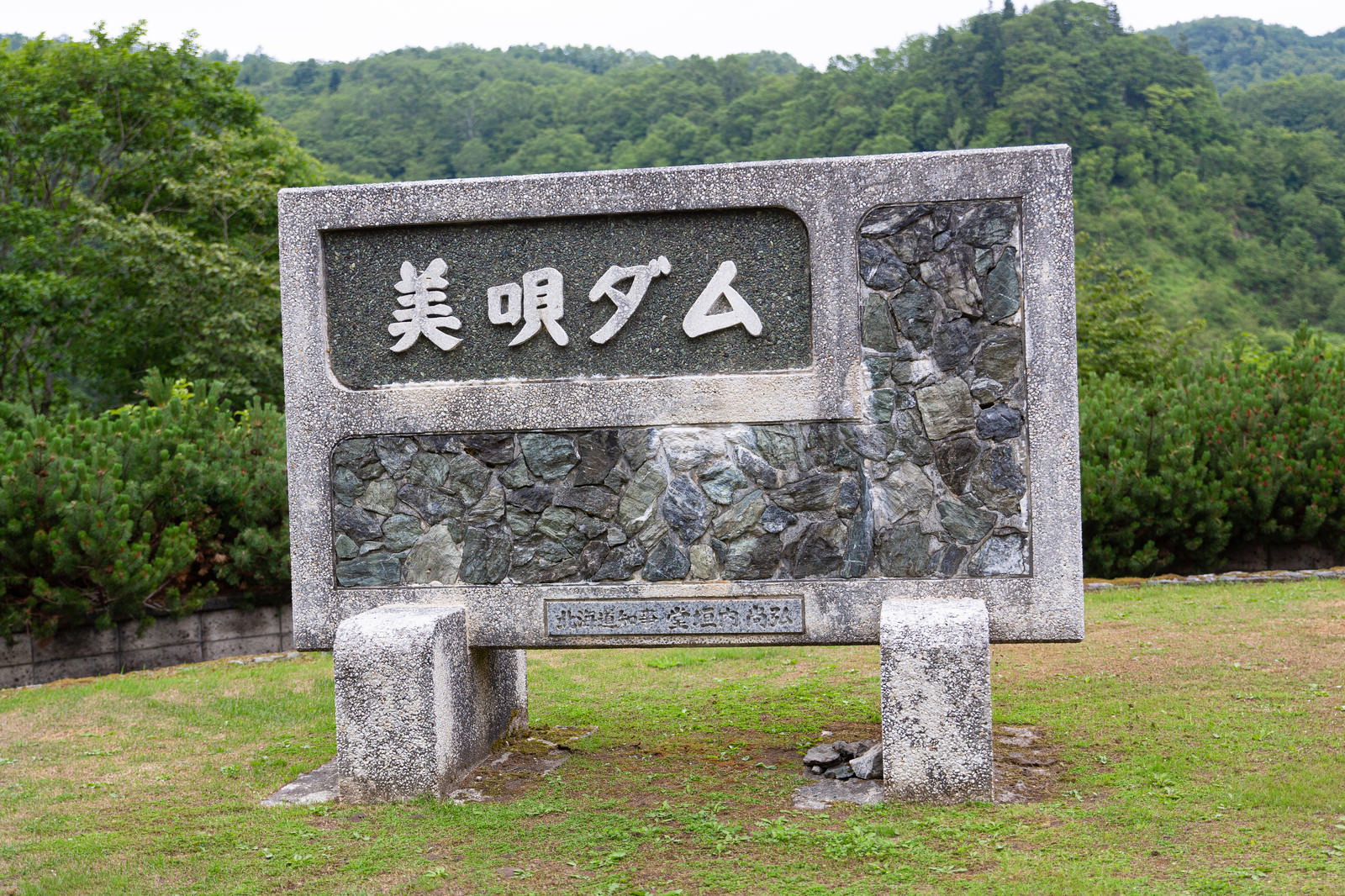 緑に囲まれた美唄ダムの石製案内板と背景の自然風景