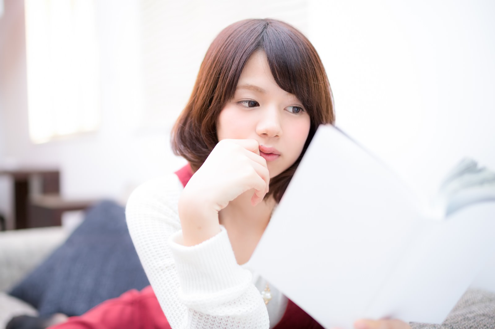 ソファーで本を読む女性が、口元に手を当てて呆れたような表情を浮かべている［モデル：みき。］