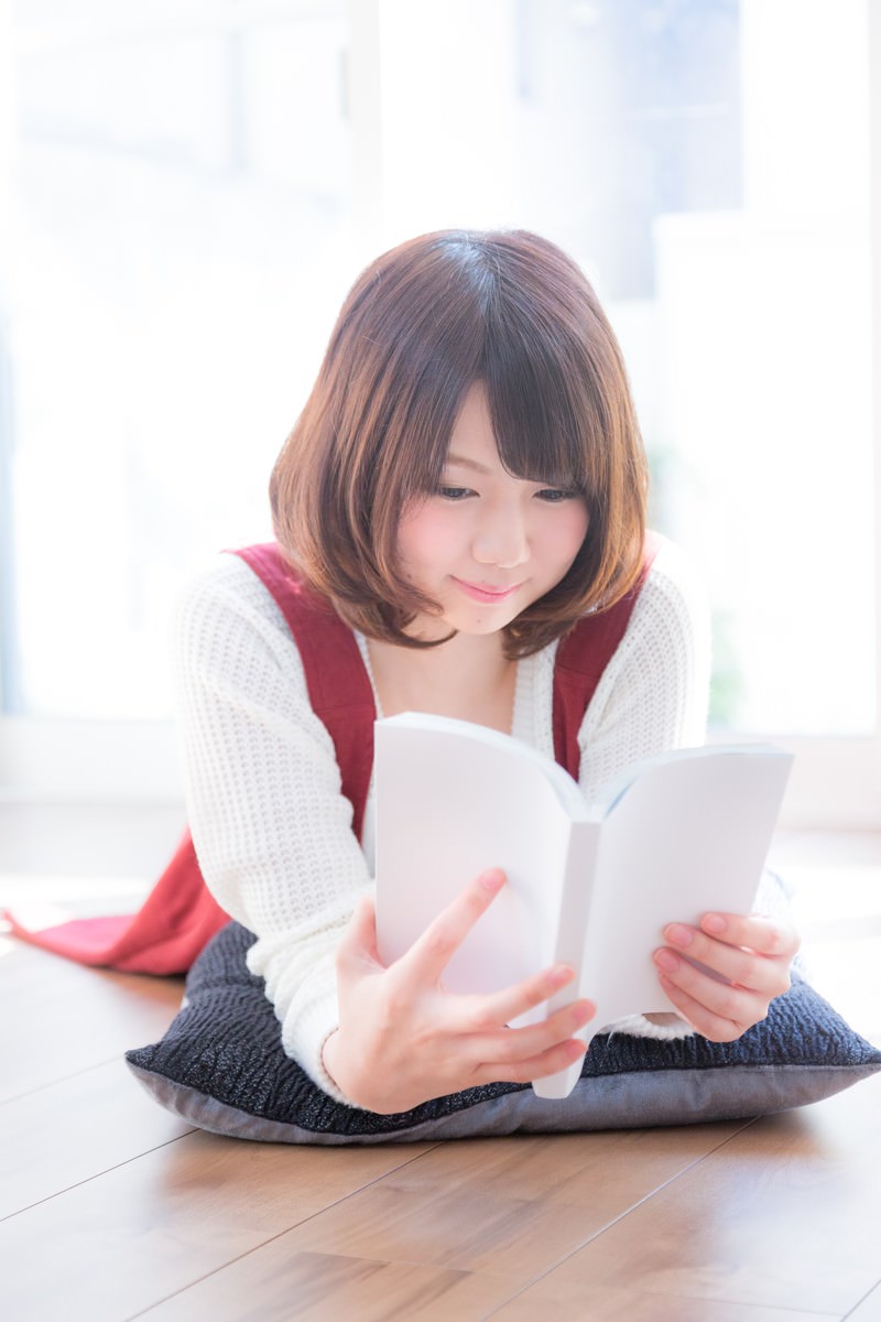 床に座ってマンガを読む赤いベストの女性［モデル：みき。］