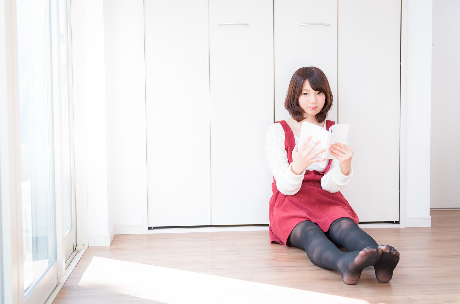 白いカーディガンと赤いスカートを着た女性がリビングの床に座ってマンガを読んでいる［モデル：みき。］