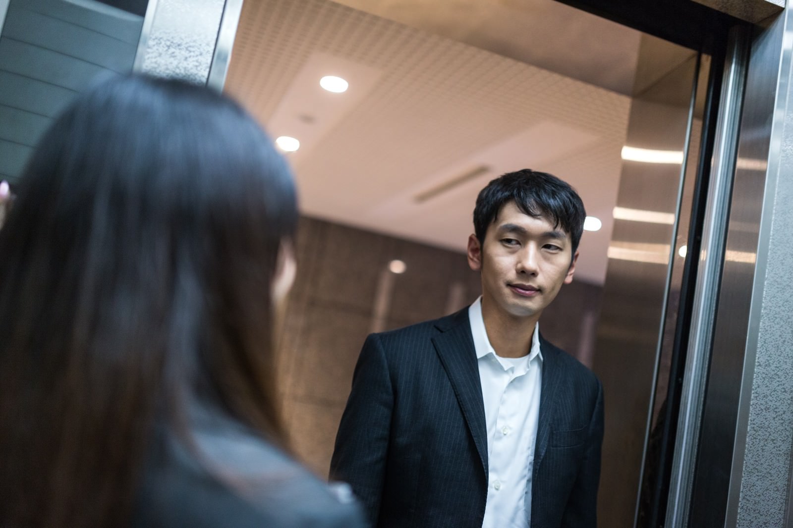 ネイビースーツの男性がうつむいた表情でエレベーター内に立ち、手前に女性の後ろ姿が見える［モデル：大川竜弥 Lala］