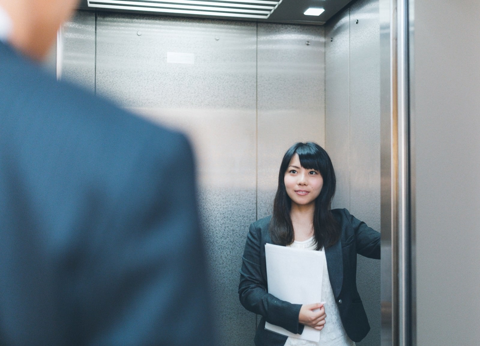 エレベーター内で来客に話しかける女性社員［モデル：Lala］