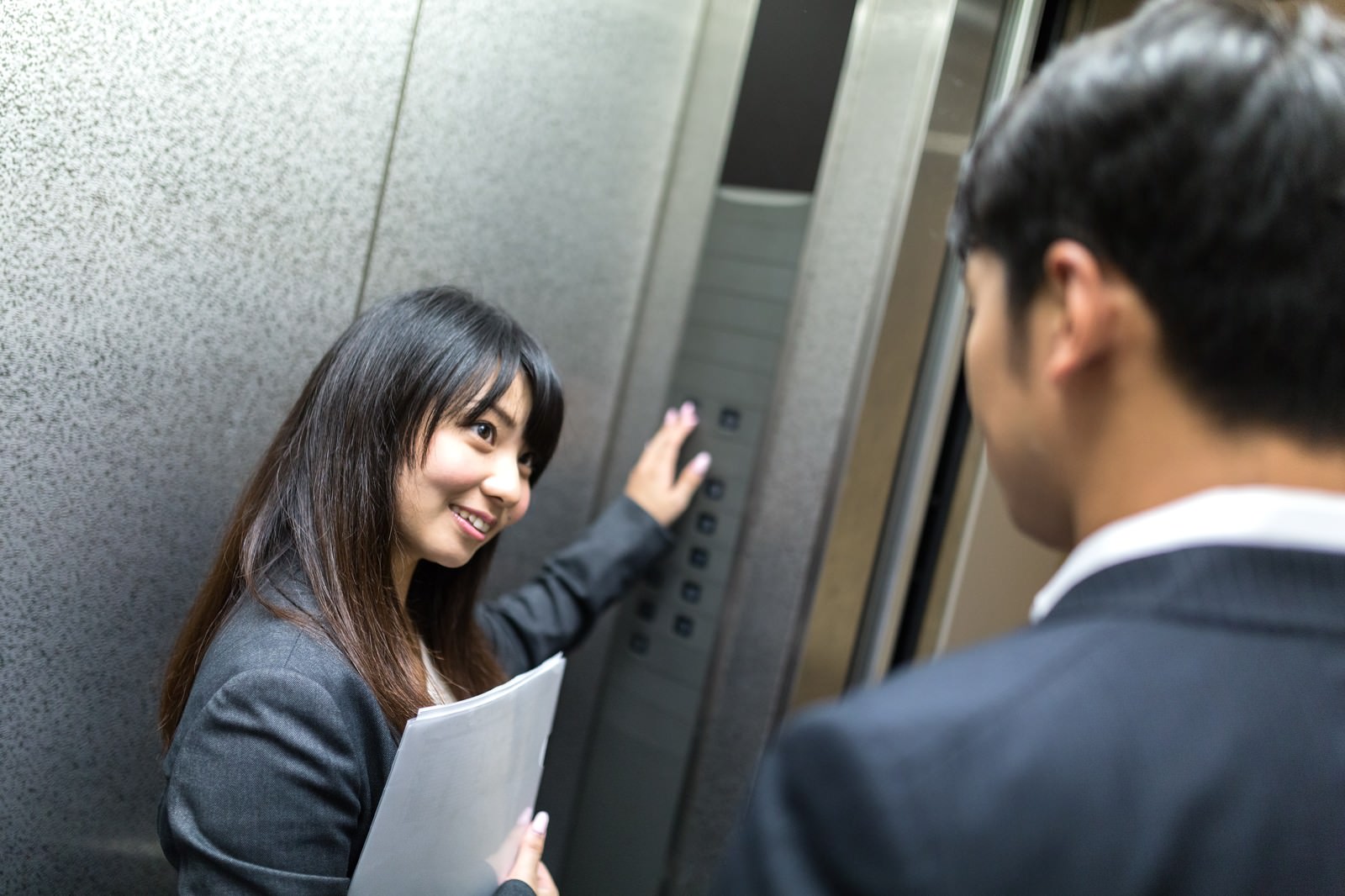 エレベーター内で振り返る黒いジャケット姿の女性と、グレースーツの男性が向き合い、二人とも笑顔でいるビジネスシーン［モデル：大川竜弥 Lala］
