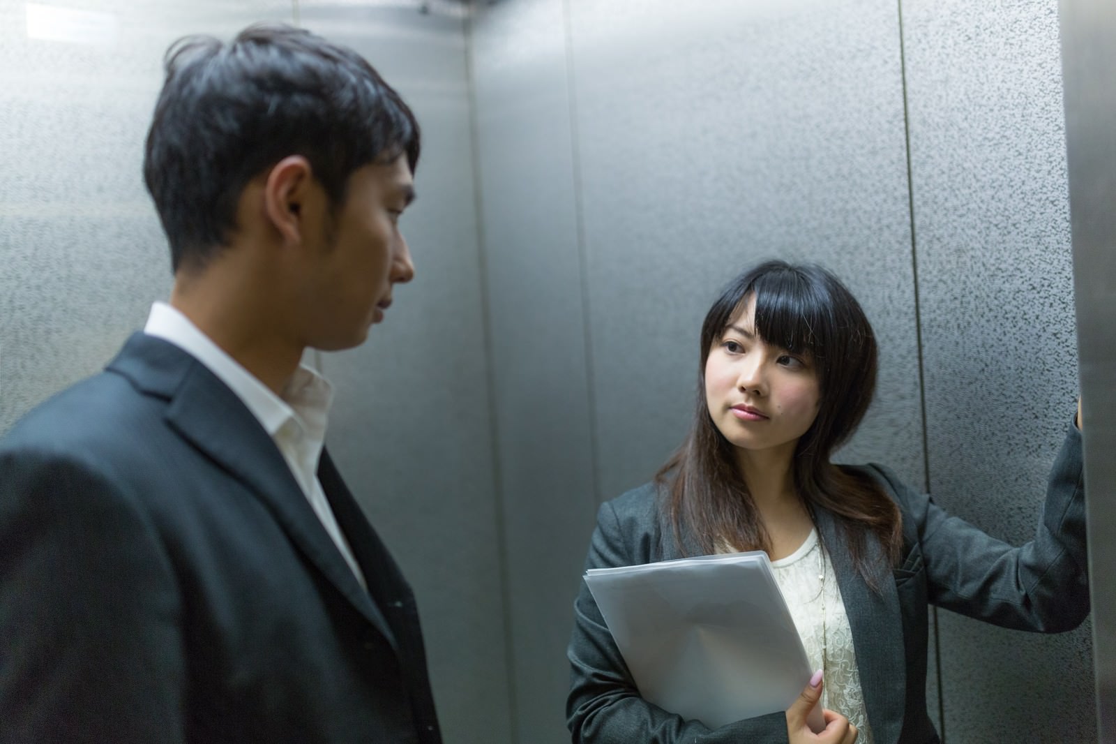 エレベーター内で紺スーツの男性に話しかけるグレースーツ姿の女性。女性はタブレットを手にしている［モデル：大川竜弥 Lala］