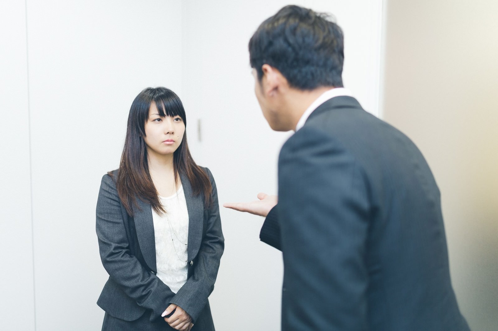 グレーのジャケットを着た女性が冷徹な表情で男性の言葉に対峙し、黒いスーツの男性が身を乗り出して訴えかけている職場シーン［モデル：大川竜弥 Lala］
