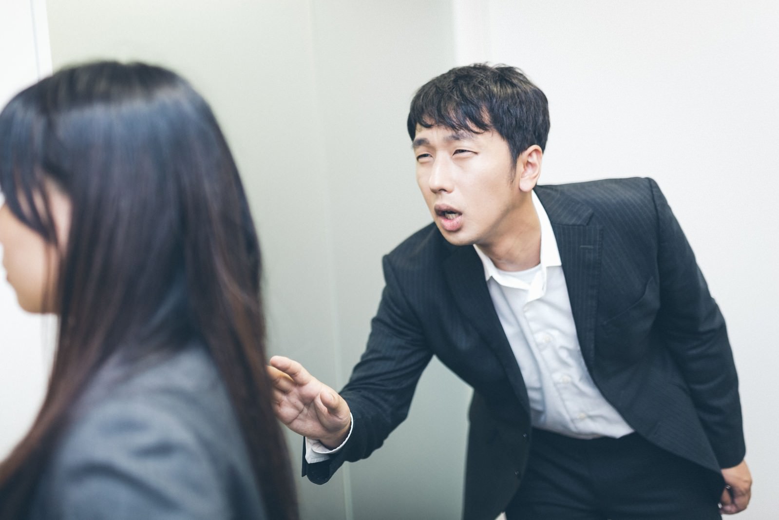 黒いスーツの男性が身を乗り出し、手を伸ばして黒髪の女性に話しかけている場面。オフィスの白い壁が背景［モデル：大川竜弥 Lala］