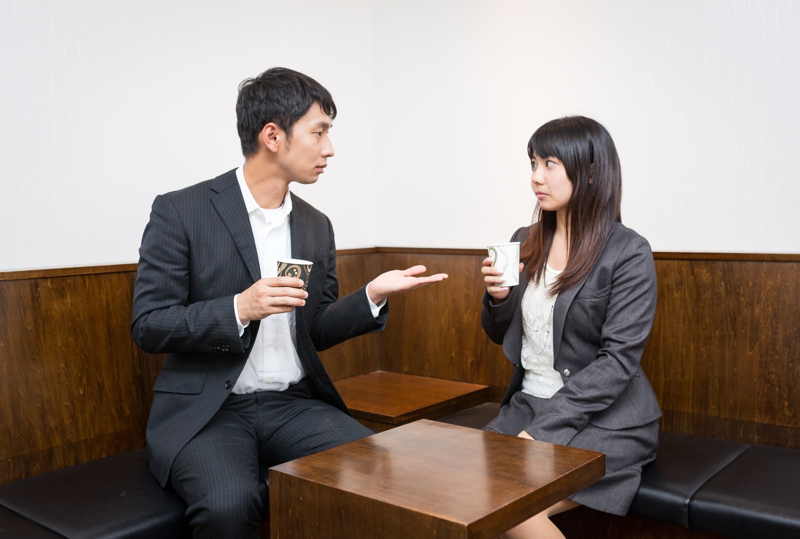 ソファに座るスーツ姿の男性と女性がカップを手に対面で会話している［モデル：大川竜弥 Lala］
