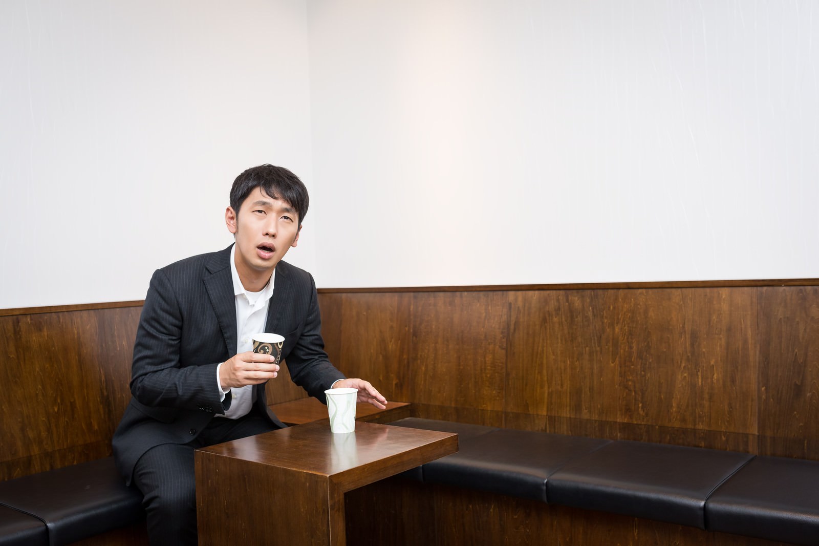 ソファーに座り紙コップを手に持ちながら上を見つめる困った表情の男性［モデル：大川竜弥］