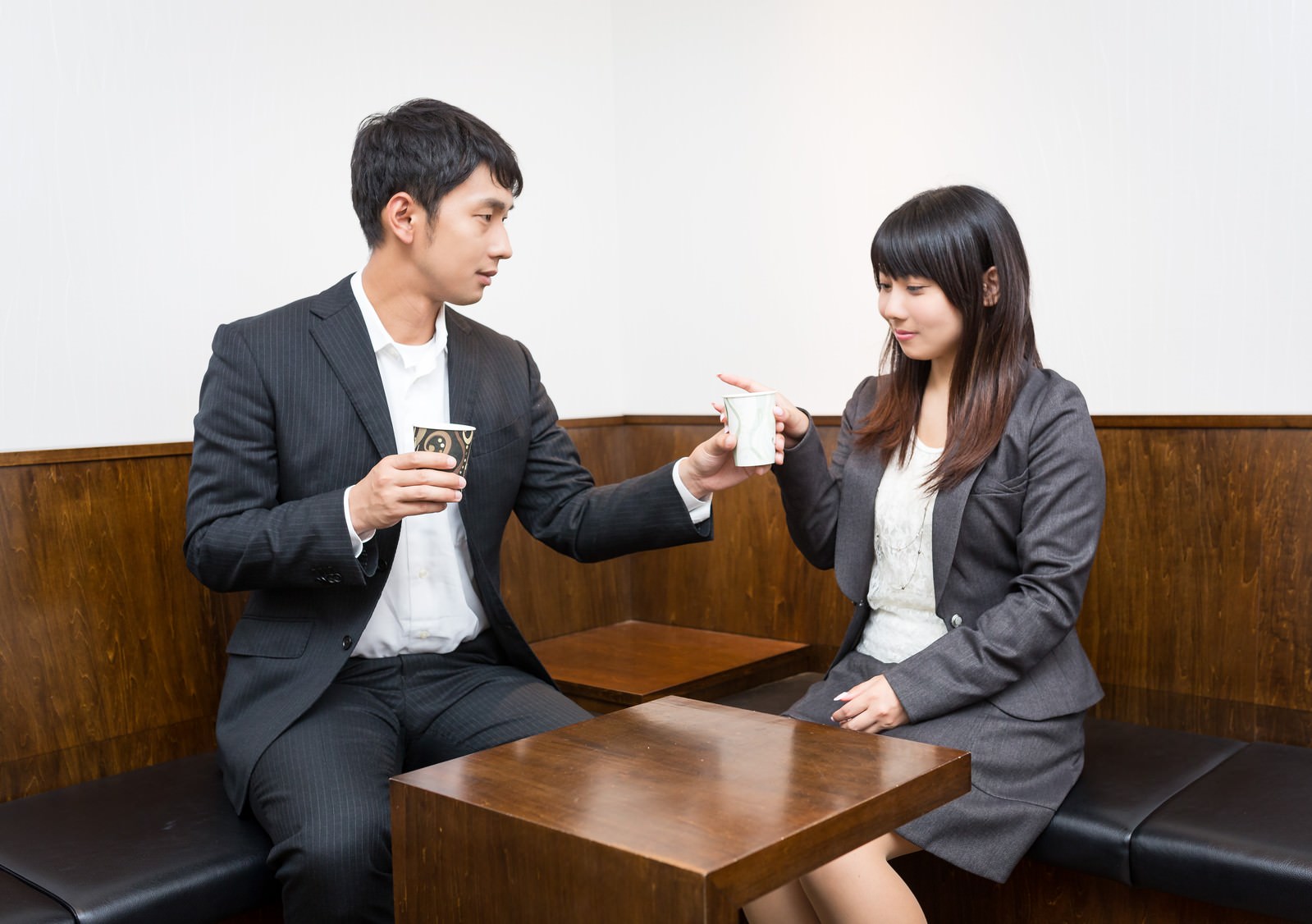 男性ビジネスパーソンが紙コップのコーヒーを女性に手渡す。木張りの受付エリアで向かい合わせで座る。［モデル：大川竜弥 Lala］
