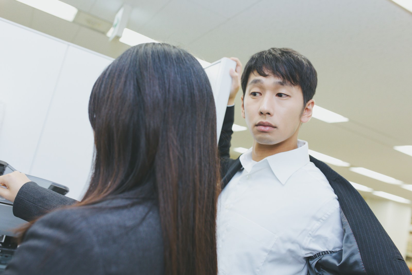 白シャツ姿の男性がジャケットを脱いでアクション中、女性社員が書類を手に持ったまま脇を通り過ぎるオフィスシーン［モデル：大川竜弥 Lala］