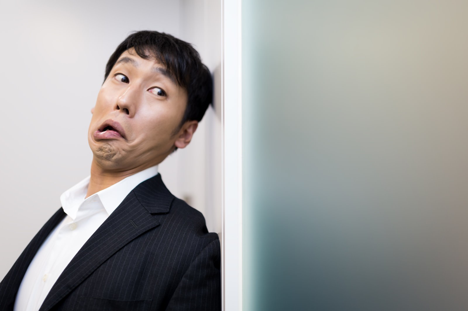 黒いスーツの男性が壁を背に驚いた表情で横目で上を見上げている［モデル：大川竜弥］