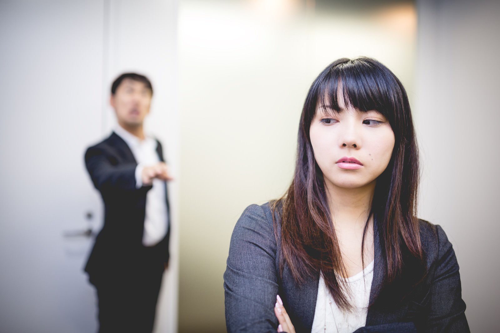 説教を受けて緊張した表情で手を握りしめる女性社員と、背後に立つ男性上司のオフィスシーン［モデル：大川竜弥 Lala］