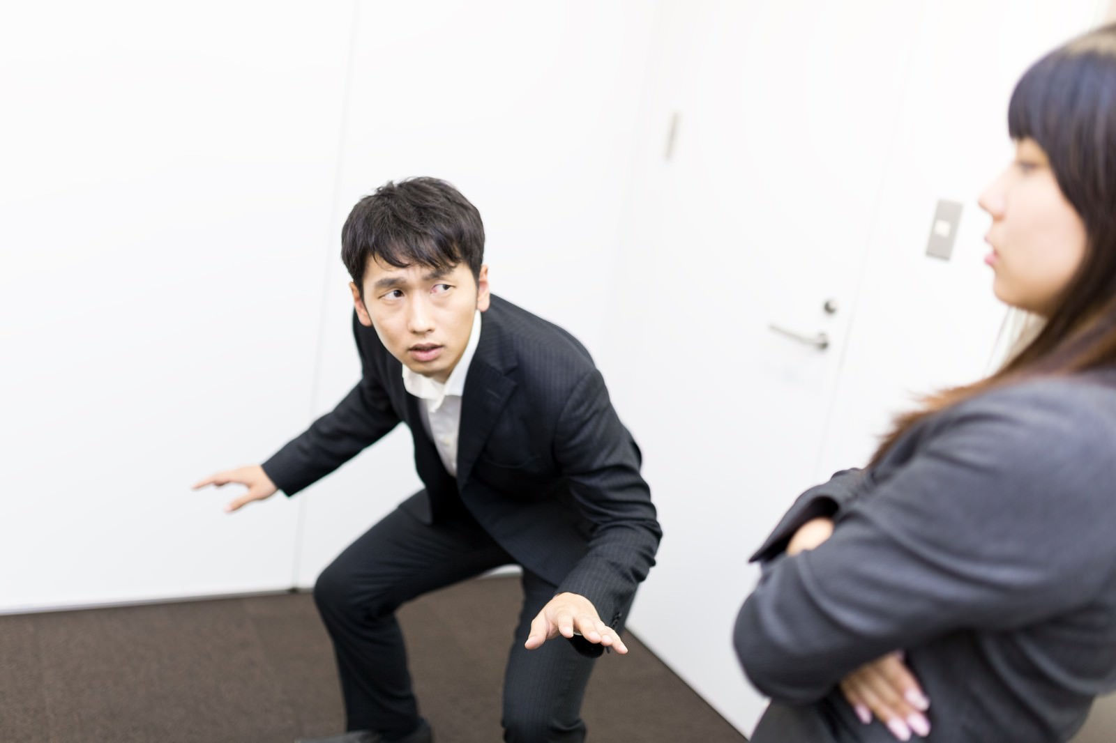スーツ姿の男性がエアサーフィンのポーズを取り、背後の女性が冷淡な表情で見つめている［モデル：大川竜弥 Lala］