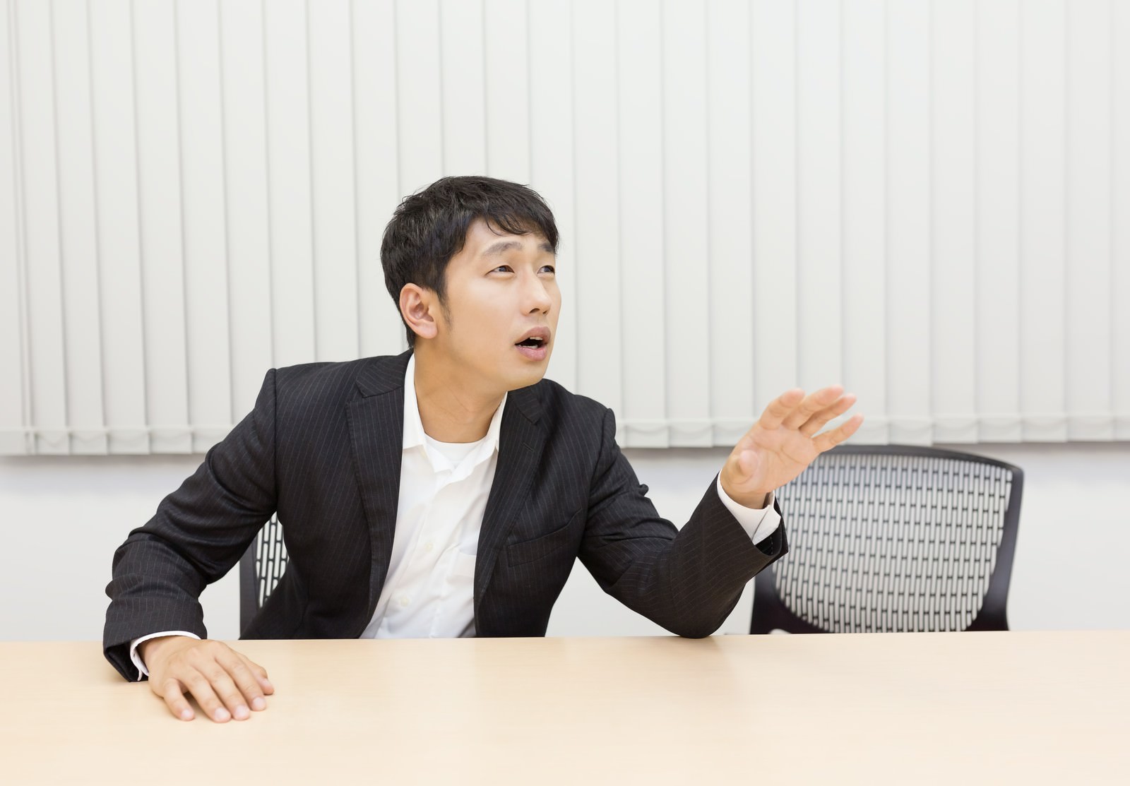 デスクで斜め上を見つめながら口を開く、躊躇した表情のビジネススーツ姿の男性［モデル：大川竜弥］