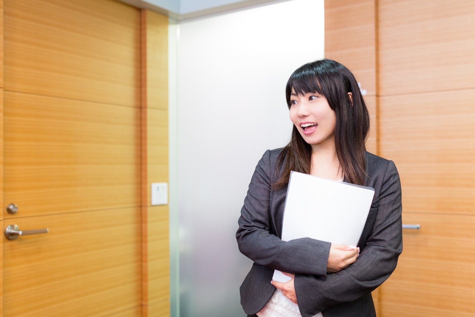 黒いスーツ姿の女性がクリップボードを抱えながら右斜め上を見つめ、明るく笑顔で挨拶している［モデル：Lala］