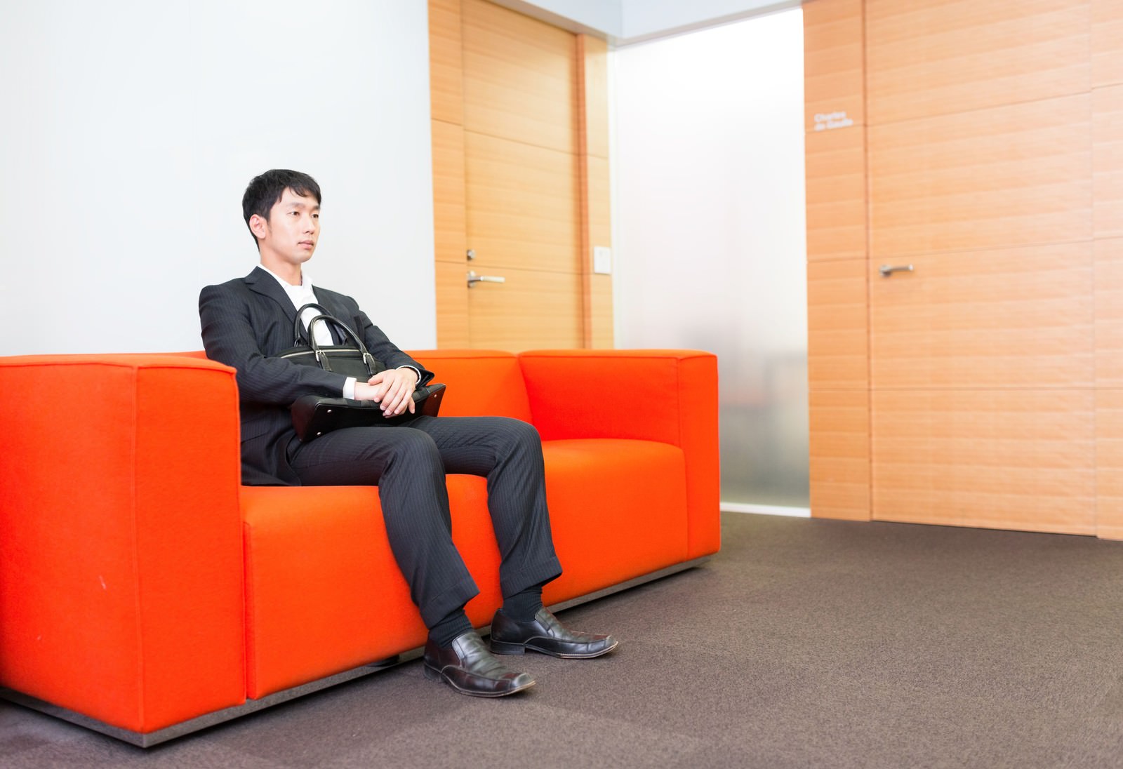 赤いソファーに座り、ハンドバッグを持つスーツ姿の男性が前を向いている［モデル：大川竜弥］