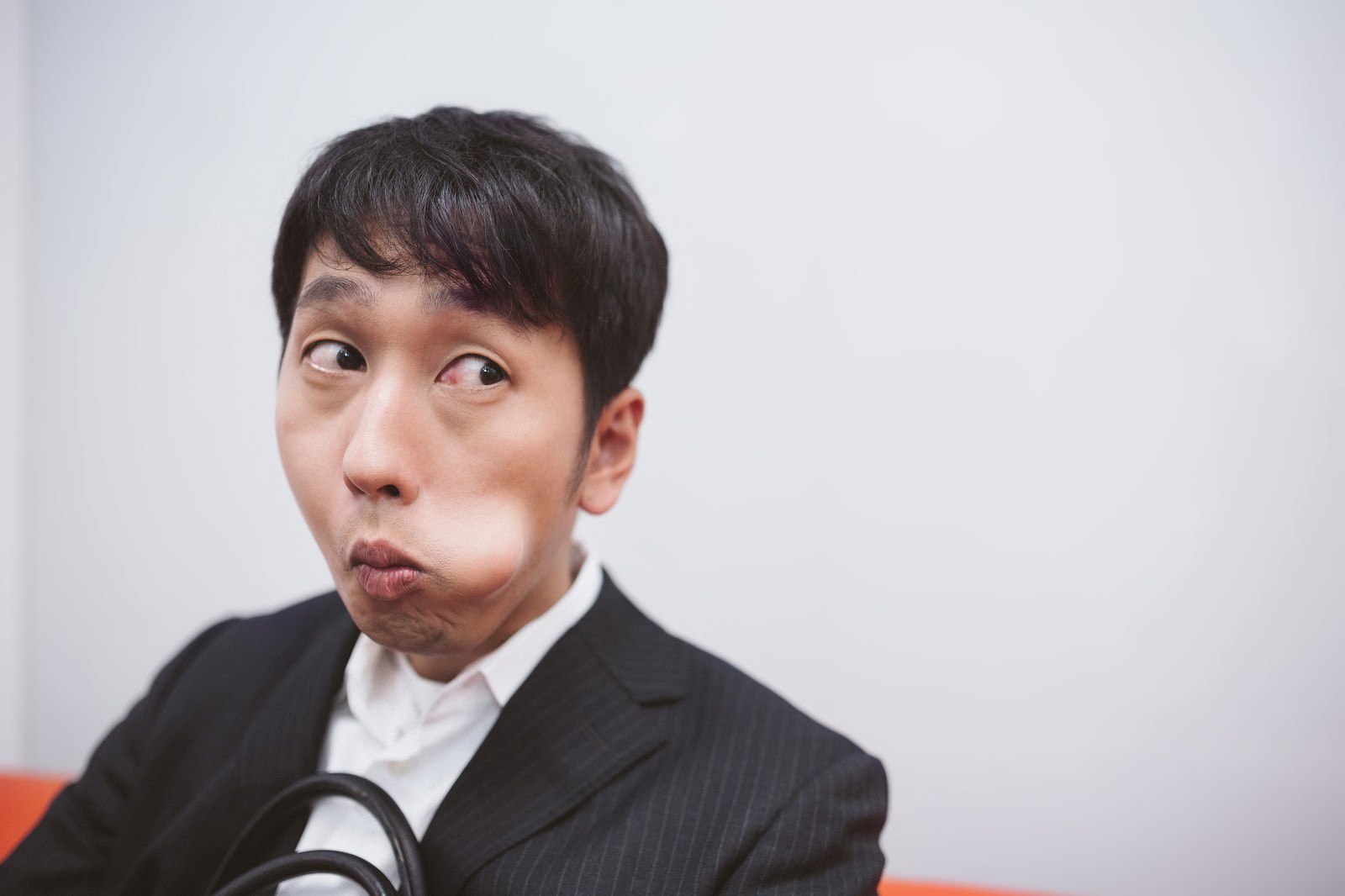 黒いスーツと白いシャツを着た男性が、目線を横に逃し頬を膨らませながら奇妙な表情で横向いている［モデル：大川竜弥］