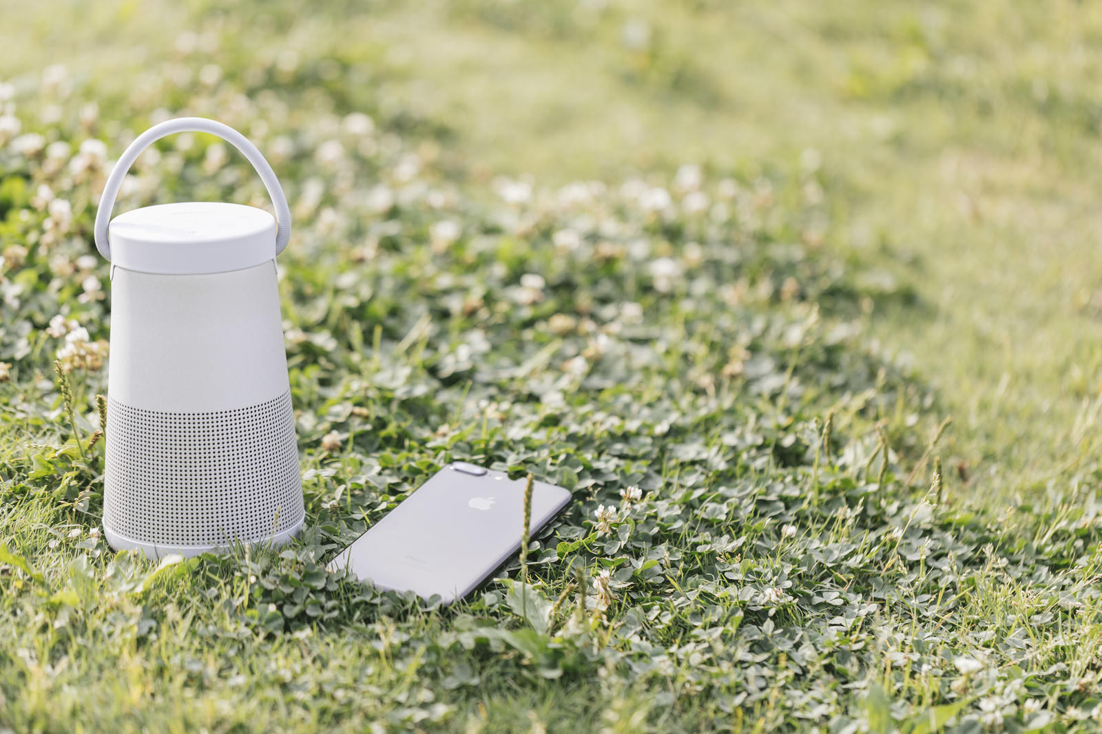 緑の芝生の上に白いBluetoothスピーカーと黒いスマートフォンが並んで置かれている