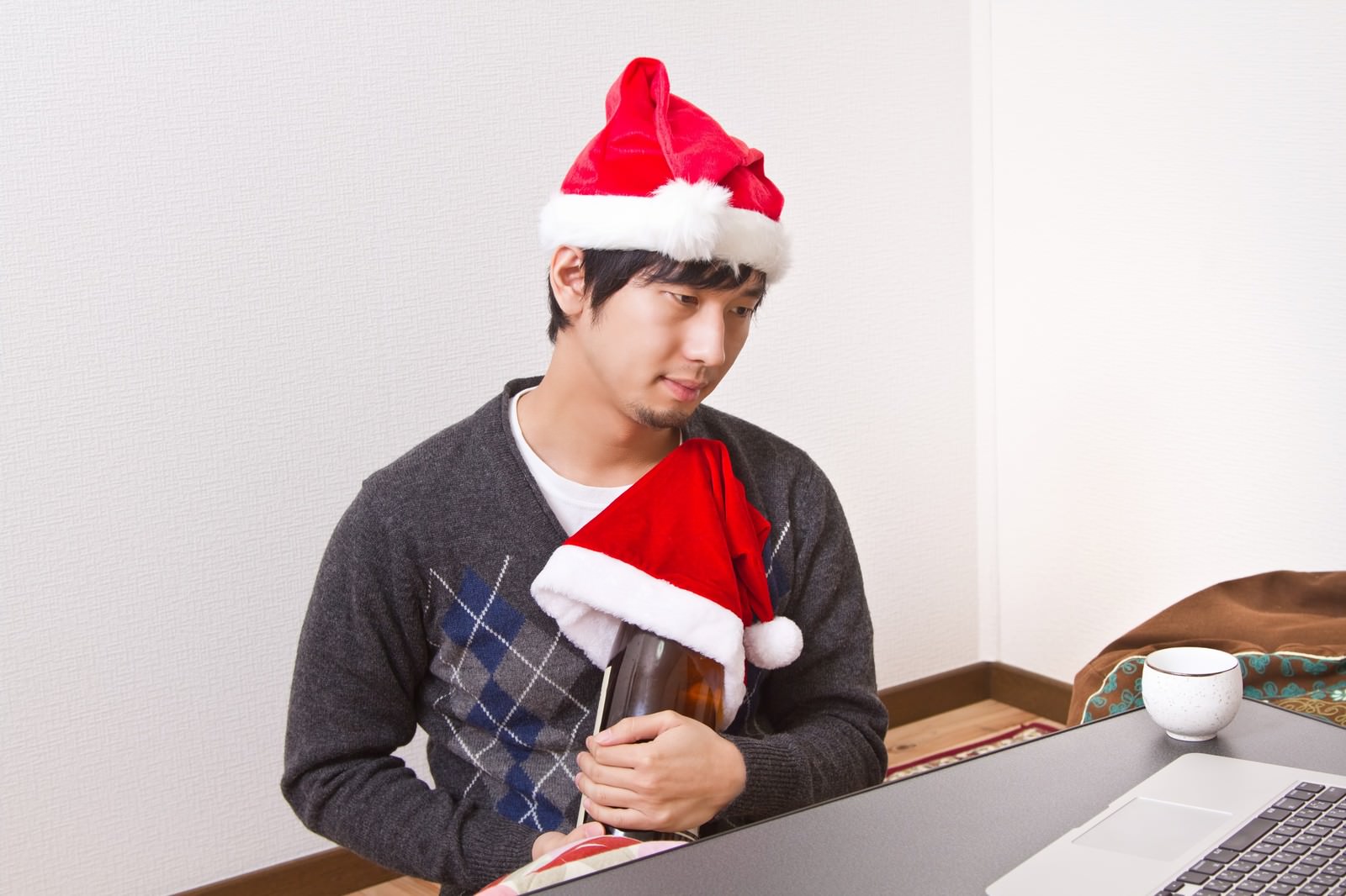 サンタ帽をかぶりPCの前で一升瓶を抱えるクリぼっちの男性［モデル：大川竜弥］
