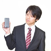 旧型のスマートフォンを手に持って画面を見るサラリーマンの写真