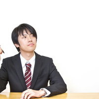 水色の壁の前でペンを持ち顎に手を当てて考えるスーツの男性の写真