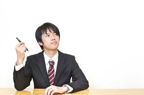 水色の壁の前でペンを持ち顎に手を当てて考えるスーツの男性