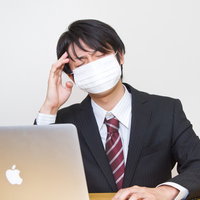 マスクを着けたままPCに向かう風邪をひいても残業するサラリーマンの写真