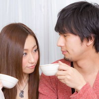 コーヒーカップを手に持ちながら笑顔で見つめ合う恋人カップルの写真