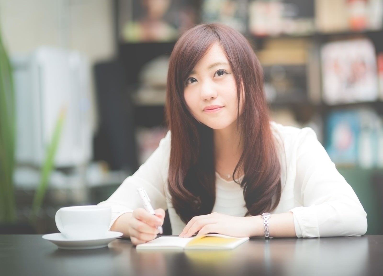 カフェのテーブルでノート開き、ペンでメモを取る女性。手前にコーヒーカップ［モデル：河村友歌］