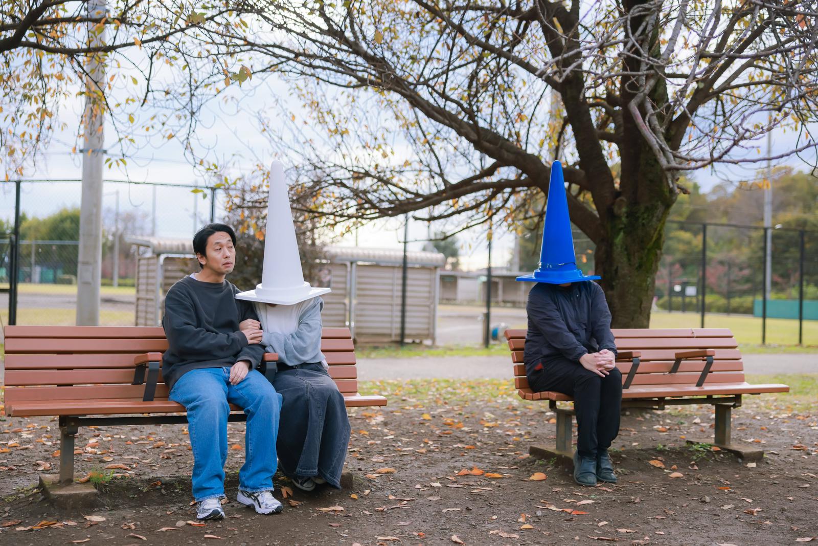 公園のベンチで白いコーン姿の人物と男性が並んで座っている［モデル：大川竜弥］