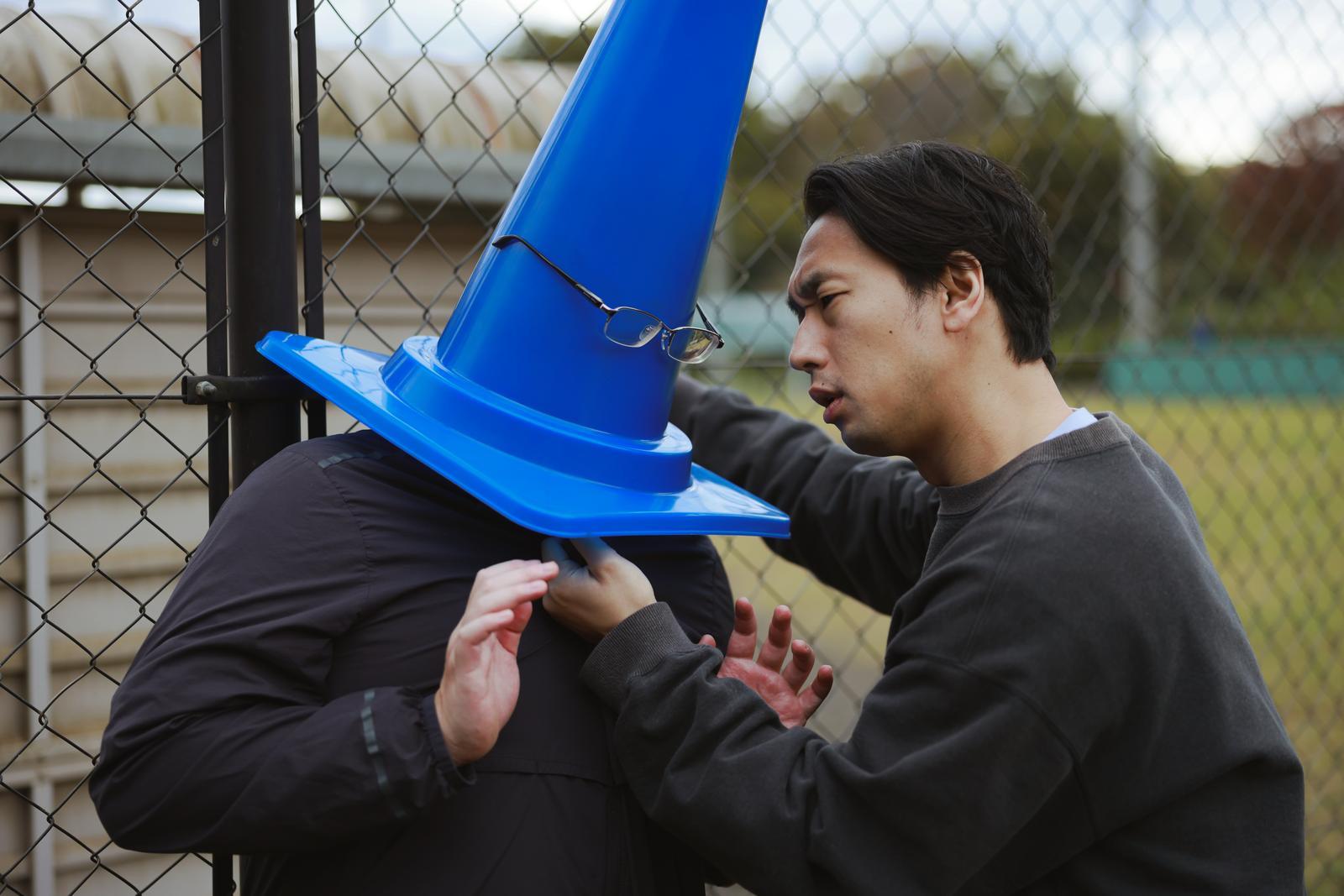 フェンス前で青い三角コーンを掴んで睨む男性［モデル：大川竜弥］
