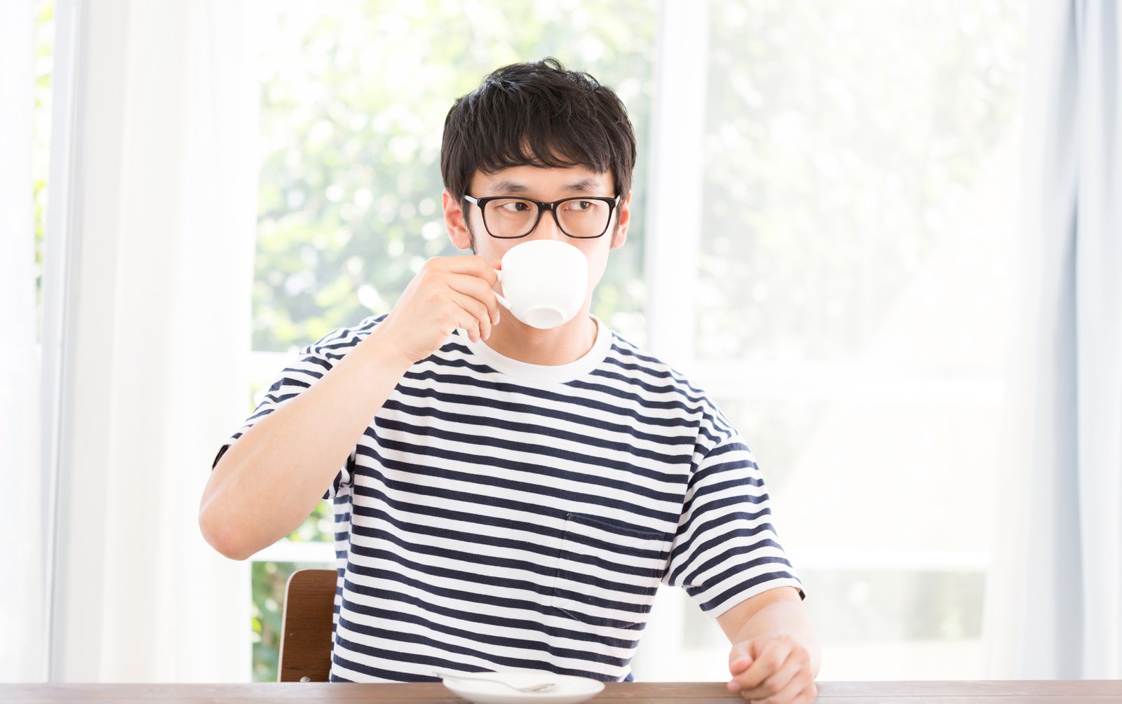 窓際で白いマグカップのコーヒーを飲む眼鏡の男性［モデル：大川竜弥］