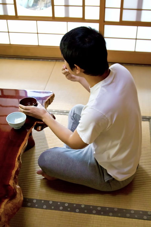 畳の和室で一人でご飯を食べる男性の後ろ姿