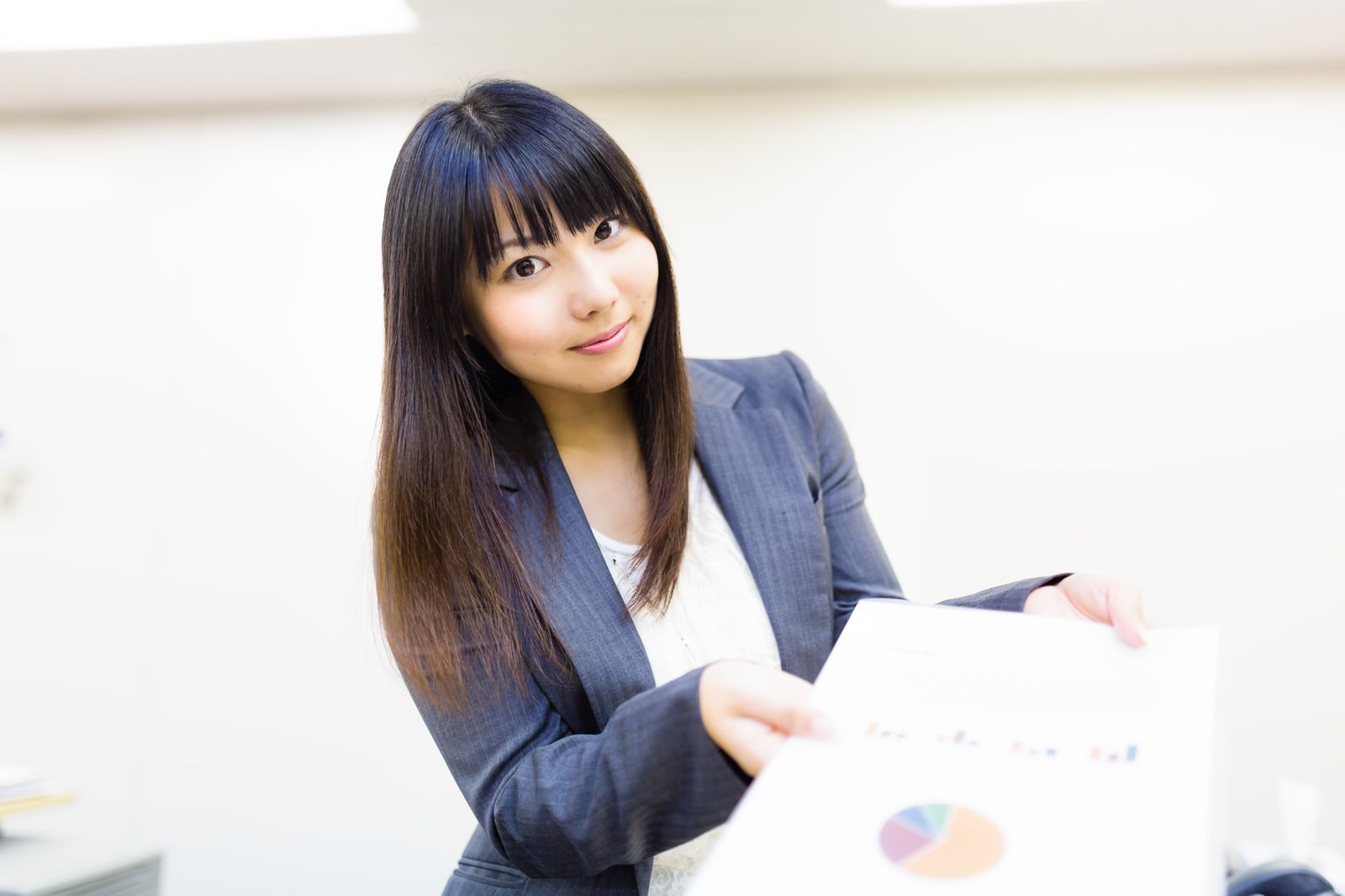 ネイビーのストライプスーツを着た女性がグラフ資料を両手で持ち、笑顔で見つめている［モデル：Lala］