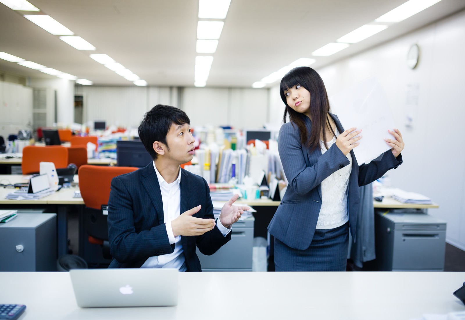 白いデスクに置かれたノートパソコンの前で、座る男性ビジネスパーソンと立つ女性が身振りを交えて書類について話し合う様子［モデル：大川竜弥 Lala］