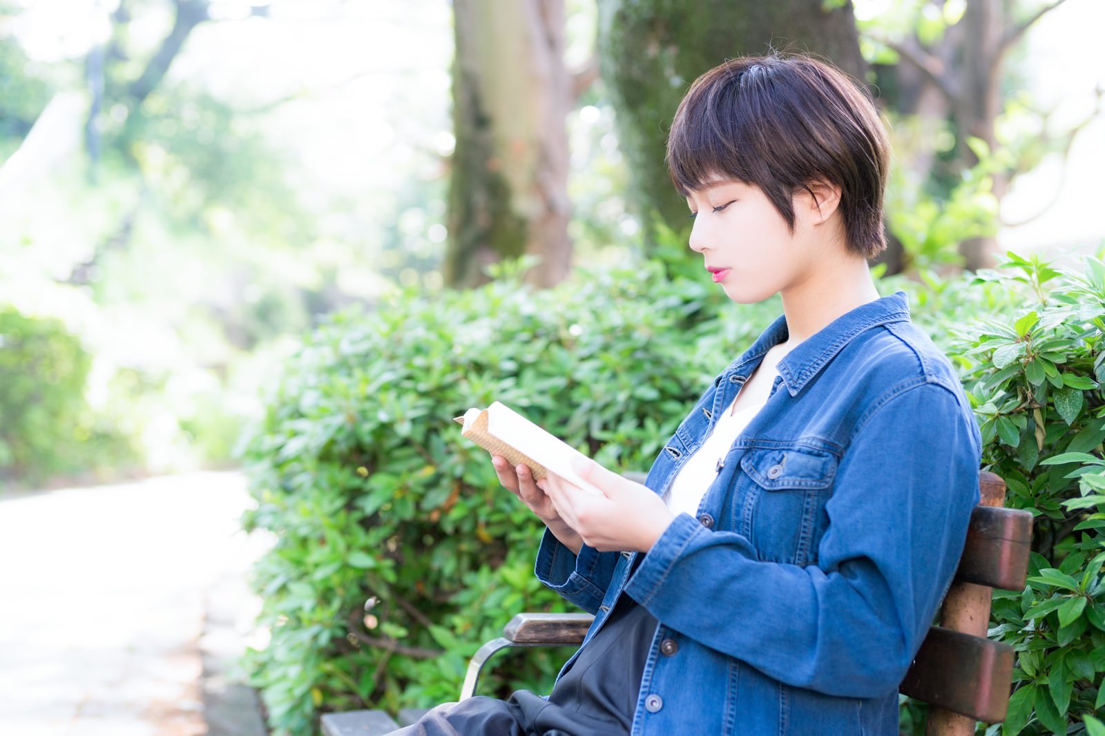 ベンチで本を読む女性。デニムシャツ姿で緑の公園で読書中［モデル：モデルヒロ］