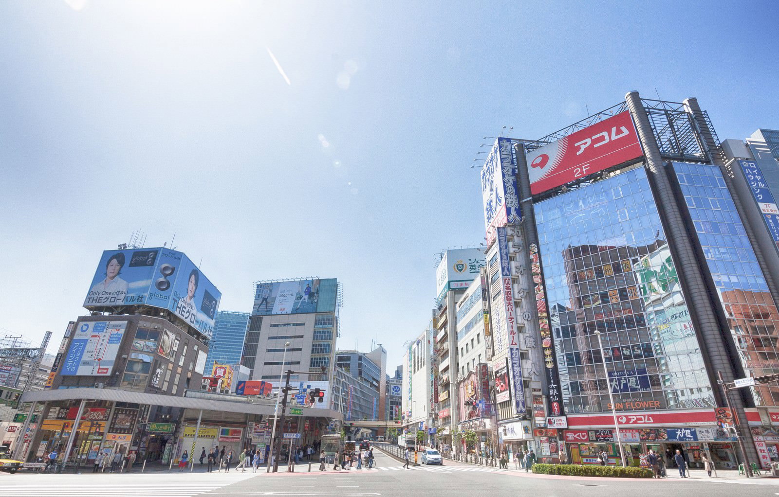 新宿駅前の青梅街道沿いで、両側に大型ビルと看板が立ち並び、多くの人が歩く繁華街の風景