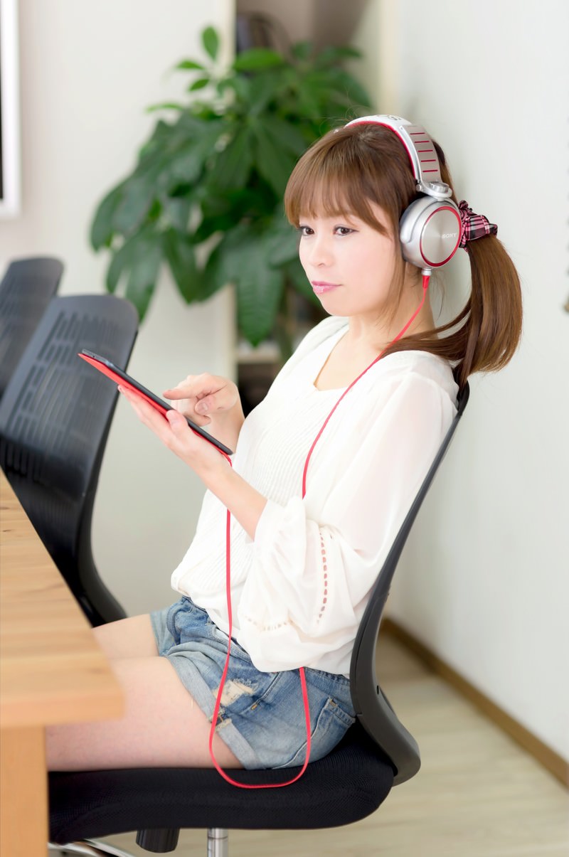 iPad miniを持ち、赤いヘッドフォンをつけてオフィスチェアに座る女性［モデル：暢子］