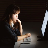 暗い部屋でモニターの光に照らされ残業する女性の写真